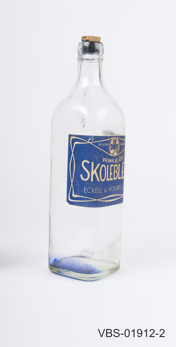 Glassflaske for skoleblekk. Firkantet gejnnomsiktig flaske med kork. 
Stor etikett på 3 sider, tekst på norsk.