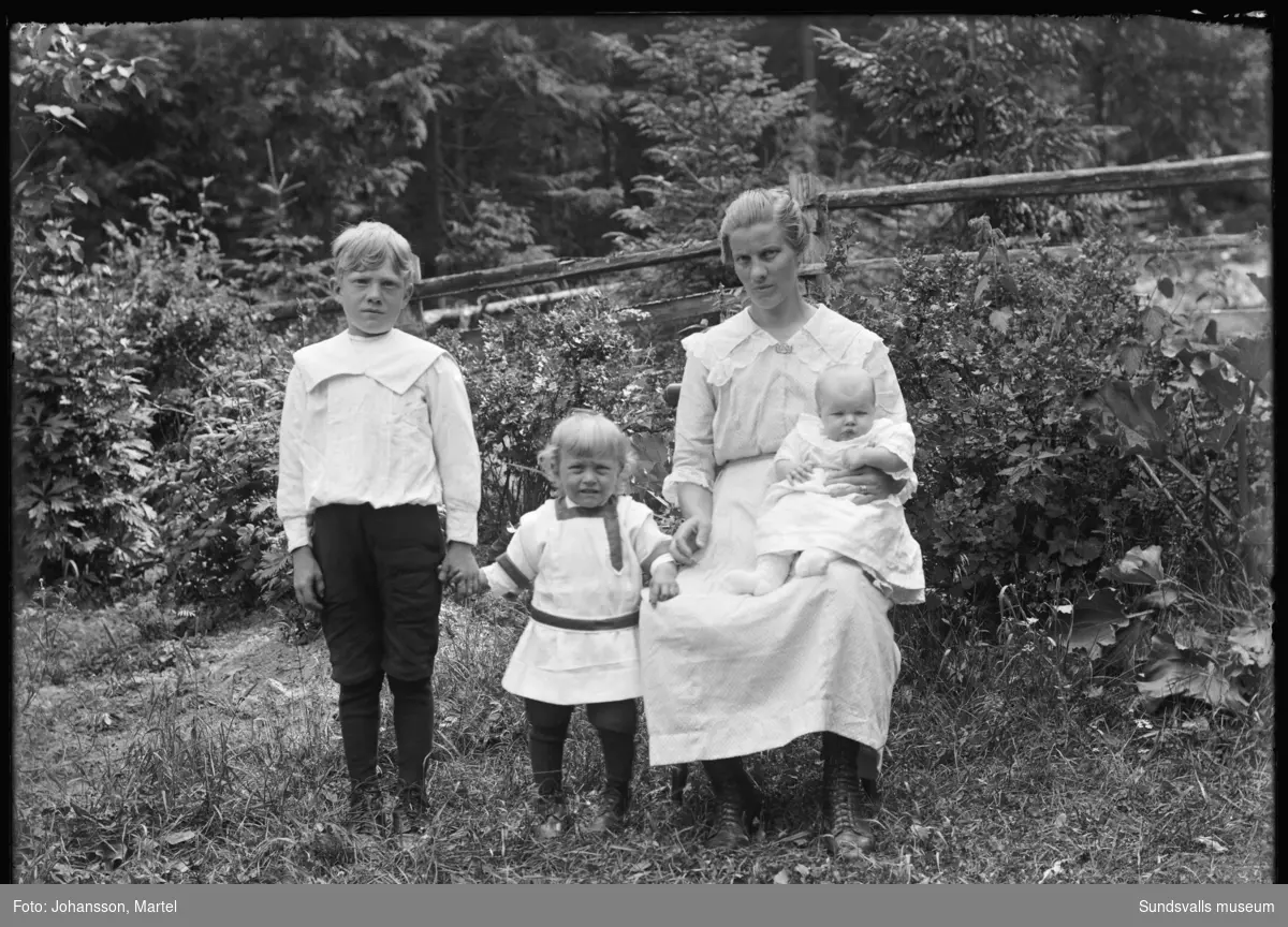 Porträtt av en kvinna med två små pojkar och en baby, fotograferat ute i en trädgård.