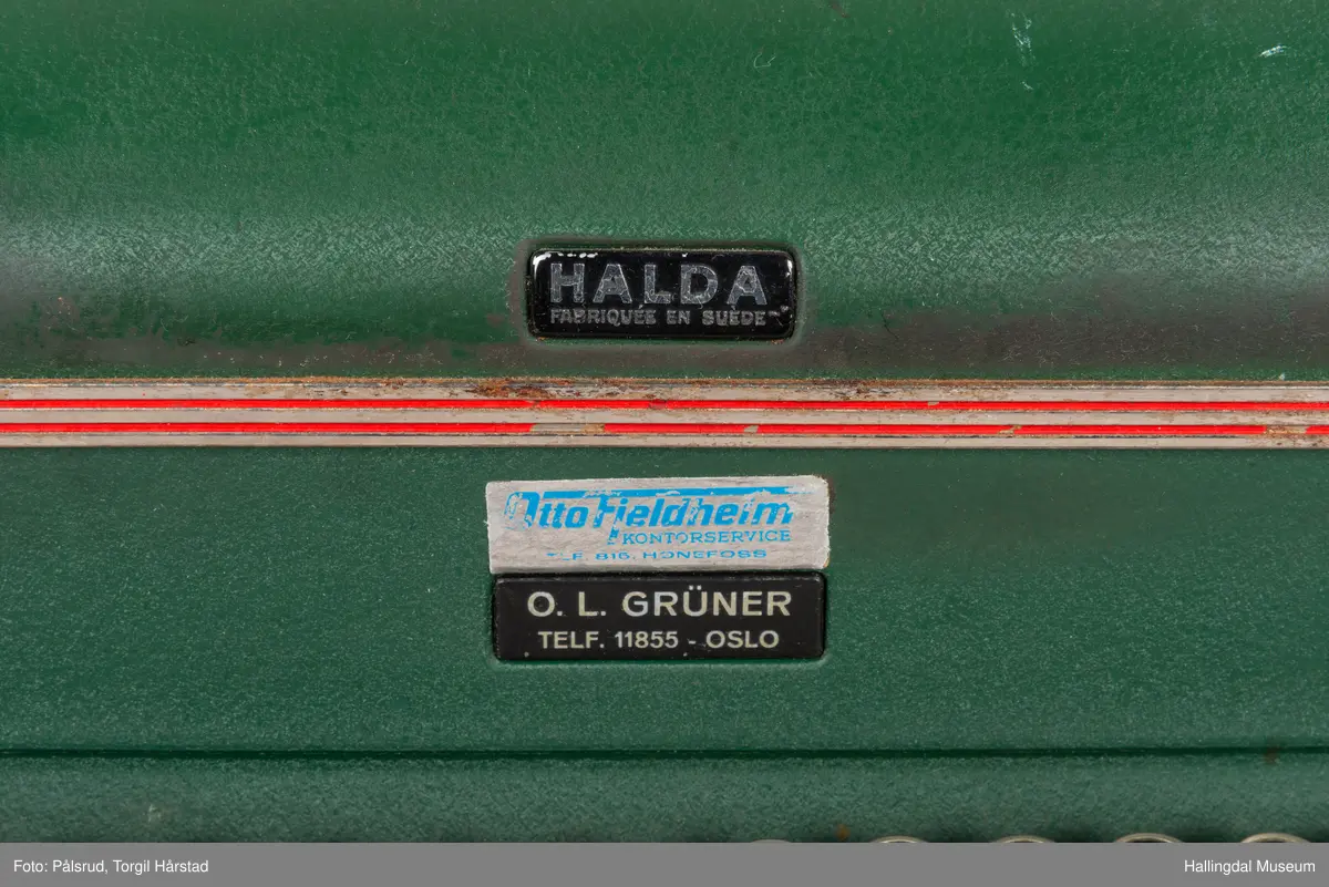 Grønn skrivemaskin av merke Halda. Den har to røde og tre gullstriper som går som en linje rundt øverste del. Noe slitasje på tastatur. 