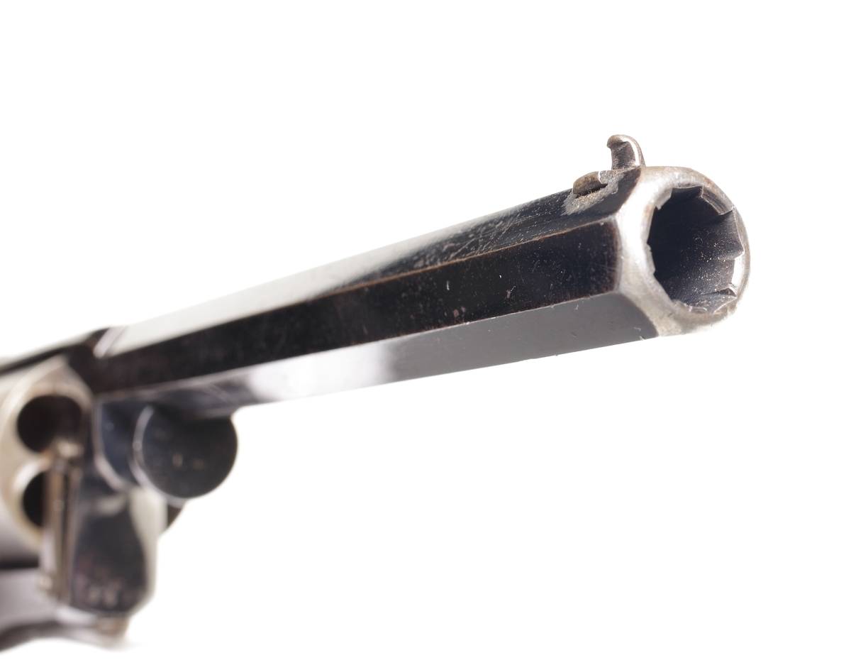 Revolver av slaglåsmodell. Den roterande trumman innehåller plats för fem laddningar. Laddning sker genom att ta loss trumman från stommen. Framför trumman sitter en spärr som trycks in, och sedan kan trumaxeln dras fram och trumman tas ur. En enkel säkring finns vid hanen som aktiveras genom att man trycker löst på avtryckaren, och samtidigt trycks säkringen in så slagstiftet inte kommer åt tändhatten. Avfyrning kan ske endast genom att trycka på avtryckaren, detta då upphakning av hanen ej är möjlig. Hela revolvern är tillverkad av blånerat stål, förutom hanen som är oblånerad. Kolvplattorna är tillverkade av mörkt ädelträ. Stomme och pipa är polerade. På kolvryggen sitter en tumplåt i silver, samt på kolvknappen en liten lucka för tändhattsförvaring. Hela vapnet är odekorerat, och pipan är åttakantig och invändigt räfflad i kaliber .54. Stomme och trumma ej nummerriktiga.
Inskrivet i huvudkatalogen 1990.