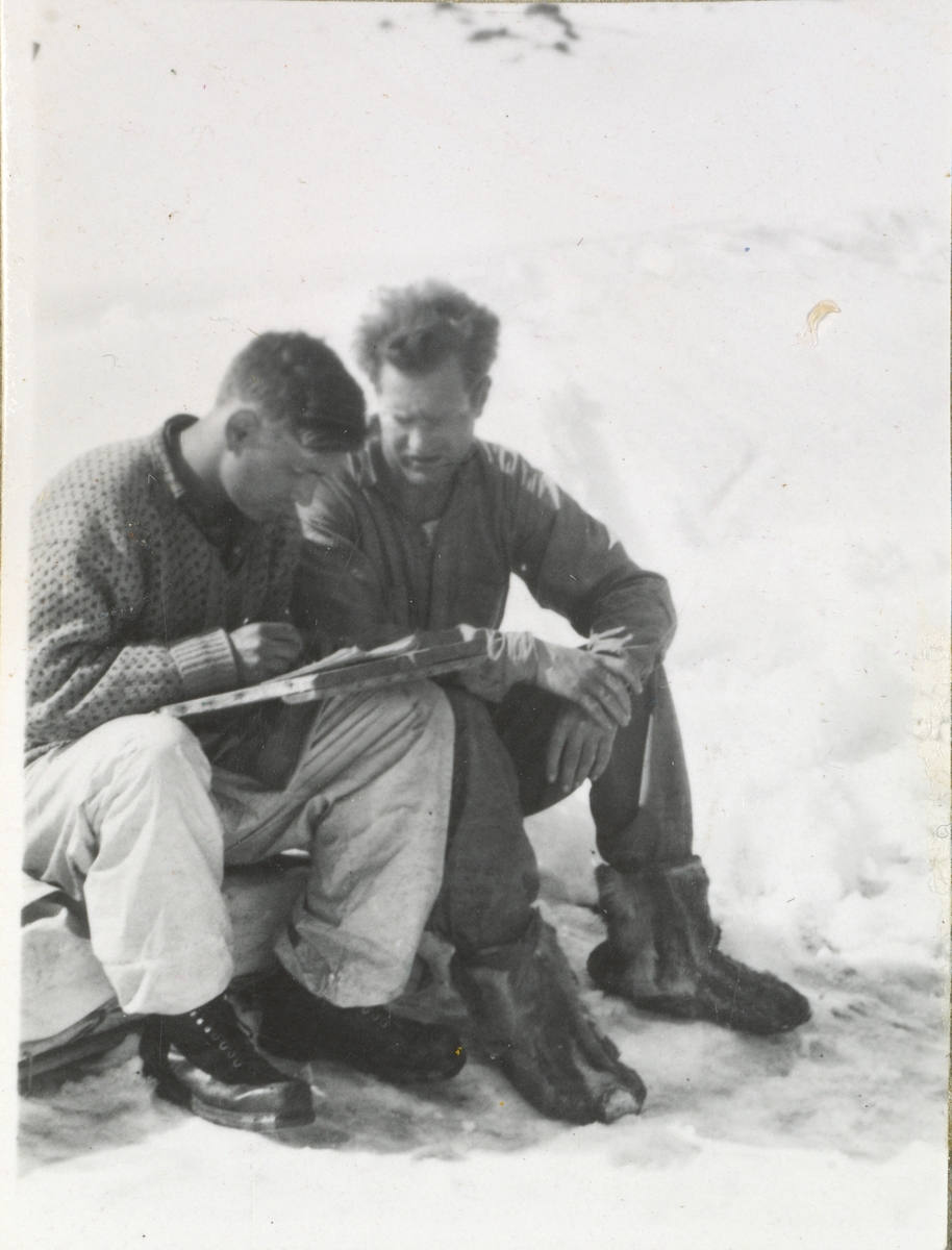 To motstandsmenn sittende i snødekket landskap i Tafjordfjellene. Olav Aarsæther har brett med papirer på fanget. Rolf Ranes har skinnsko på føttene.