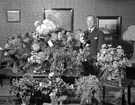 Man i hemmiljö omgiven av blommor.
Fotografens ant: Oscar Stark 50 års dag. 1935.