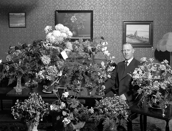Man i hemmiljö omgiven av blommor.
Fotografens ant: Oscar Stark 50 års dag. 1935.
