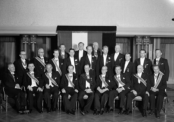 Större grupp män med ordensband.
Fotografens ant: Herr Lindqvist. Trikåfabriken.
Företaget grundades 1914 och då byggdes en fabrik upp på Herrhagen i Karlstad. I början tillverkades enbart bomullstrikå men efter några år togs även ylle och silke upp i produktionen samtidigt som fabriken utökades med eget färgeri och blekeri. 1937 inköpte bolaget Dala Väveri AB, Tidafors och Molkom som drevs som under namnet AB Värmlands Trikåfabrik avdelning B. Fabrikerna var mycket moderna med bland annat glastak för att tillvarata dagsljuset och högtalaranläggningar för att genom musik ge de anställda stimulans i arbetet.
Källa: Nyblom-Svanqvist, Näringsliv i Värmland, 1945.
Namnuppgifter saknas. Text på negativkuvert: Herr Lindqvist. Trikåfabriken.