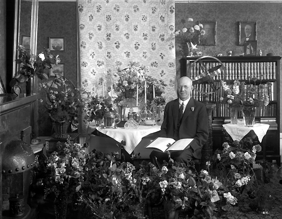 Man med bok i handen omgiven av blomsterarrangemang.
Fotografens ant: Direktör Forsgren.