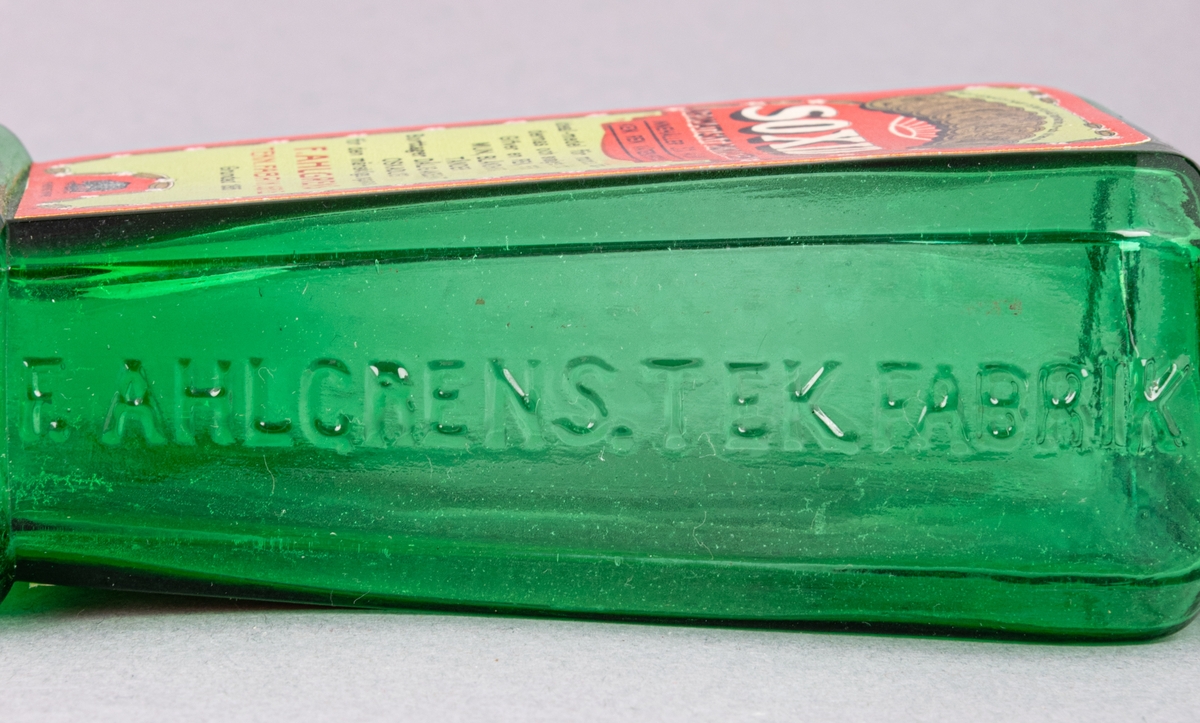 Glasflaska för munvatten, grön, rektangular, med kork. Profilerad skuldra. Etiketter på fram- och baksida. Präglad text på sidorna.