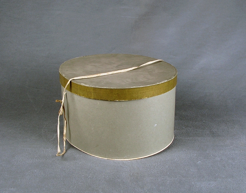 Hattaskt av ljusgrå papp. a) ask, b) lock. Oval form och i locket ett bomullsband att bära i. Locket är kantat av grågrönt papper som avviker lite i färgton från övriga asken och locket. Både asken och locket är märkta med en stämplad åtta i botten.