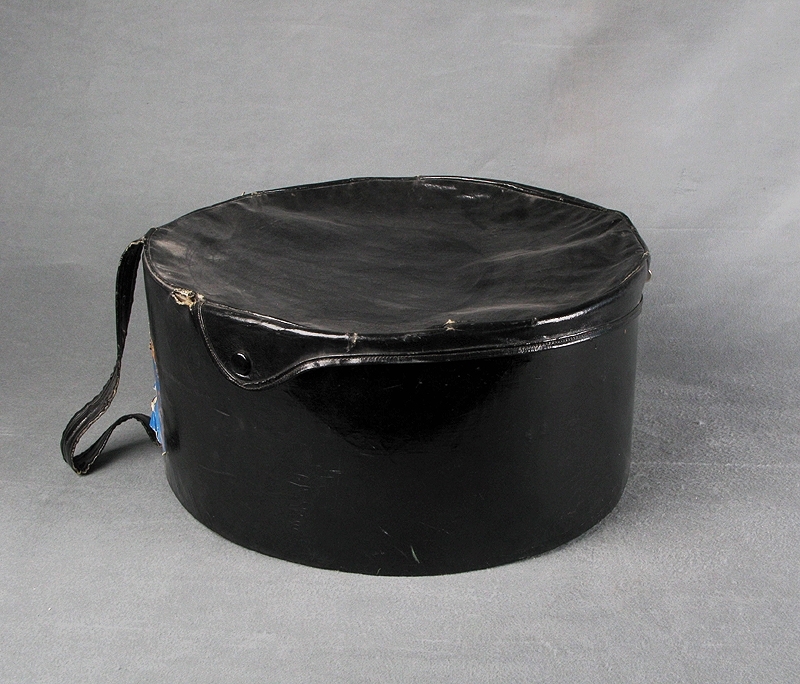 Hattask eller hattväska av svart galonmaterial stomme av papp och insida fodrad med tyg i abstrakt mönster i orange, lila och svart. I botten på insidan finns lila bomullsband att förankra hatten i. Locket sitter ihop med asken med en söm i ungefär en tredjedel av askens överkant. Resten av locket tillsluts med ett par tryckknappar och ett vred när asken är stängd. Handtag av svart galon att bära väskan i. Märkt med blå klisterlapp. Text i guld: "M/N SORRENTO SOC. PARTENOPEA DI NAVIGAZIONE"