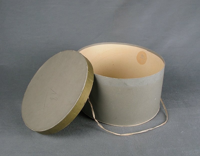 Hattaskt av ljusgrå papp. a) ask, b) lock. Oval form och i locket ett bomullsband att bära i. Locket är kantat av grågrönt papper som avviker lite i färgton från övriga asken och locket. Både asken och locket är märkta med en stämplad åtta i botten.