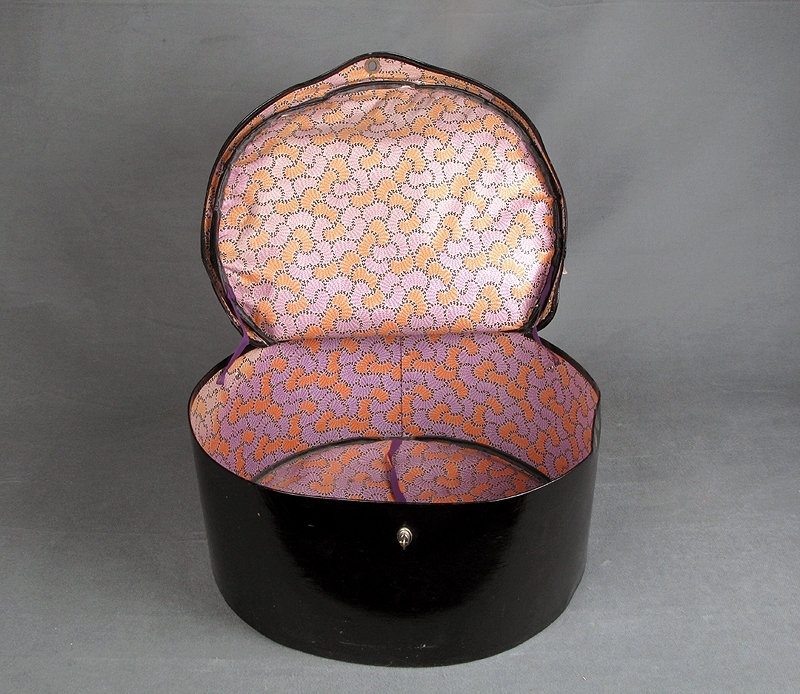 Hattask eller hattväska av svart galonmaterial stomme av papp och insida fodrad med tyg i abstrakt mönster i orange, lila och svart. I botten på insidan finns lila bomullsband att förankra hatten i. Locket sitter ihop med asken med en söm i ungefär en tredjedel av askens överkant. Resten av locket tillsluts med ett par tryckknappar och ett vred när asken är stängd. Handtag av svart galon att bära väskan i. Märkt med blå klisterlapp. Text i guld: "M/N SORRENTO SOC. PARTENOPEA DI NAVIGAZIONE"