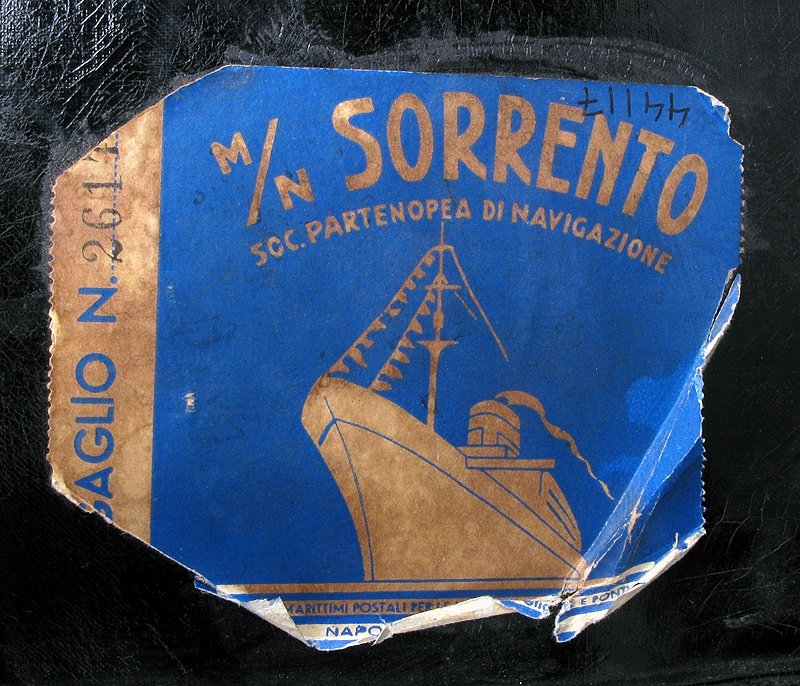 Hattask eller hattväska av svart galonmaterial stomme av papp och insida fodrad med tyg i abstrakt mönster i orange, lila och svart. I botten på insidan finns lila bomullsband att förankra hatten i. Locket sitter ihop med asken med en söm i ungefär en tredjedel av askens överkant. Resten av locket tillsluts med ett par tryckknappar och ett vred när asken är stängd. Handtag av svart galon att bära väskan i. Märkt med blå klisterlapp. Text i guld: "M/N SORRENTO SOC. PARTENOPEA DI NAVIGAZIONE"