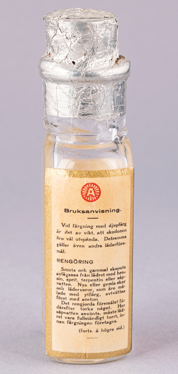 Rektangulär glasflaska för skofärg, med kork. Röd etikett med tryck i svart och vitt. Bruksanvisning på sidan.