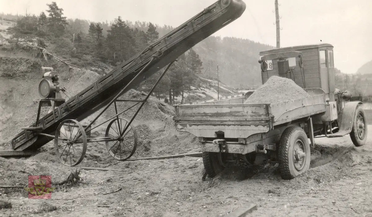 Grustransport. Bilen er en GMC lastebil, antakelig 1926 modell.
Transportbåndet er en tidlig modell med motordrift.
Bildet er fra grustak i Oftedal i Lyngdal.