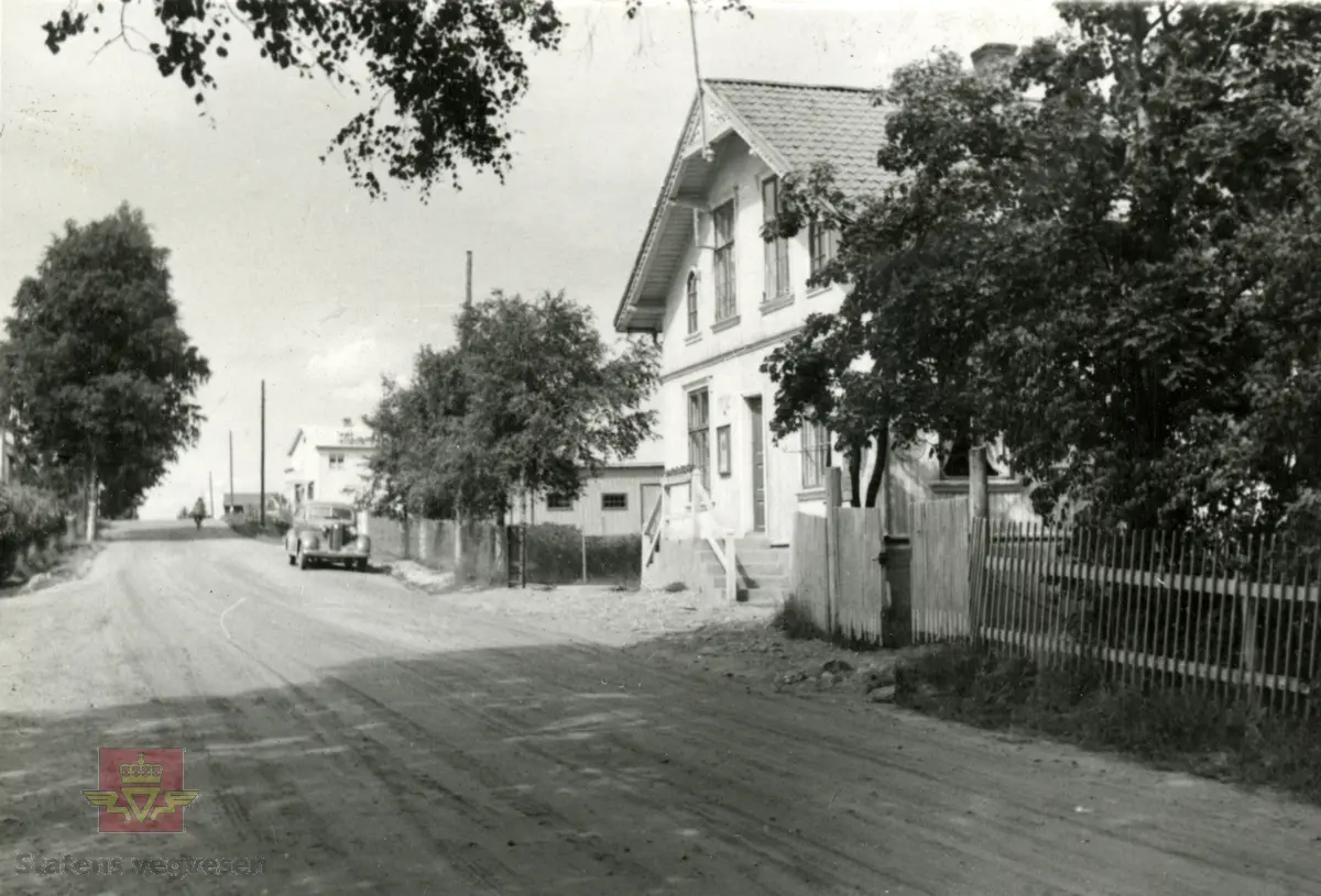 Riksveg 101 Magnor D11 i følge merking i fotoperm. Bildet viser vegen som går langs et område med bolighus  i nærheten av Magnor stasjon. En bil står parkert. Bildet er trolig tatt i begynnelsen  av 1950-årene.