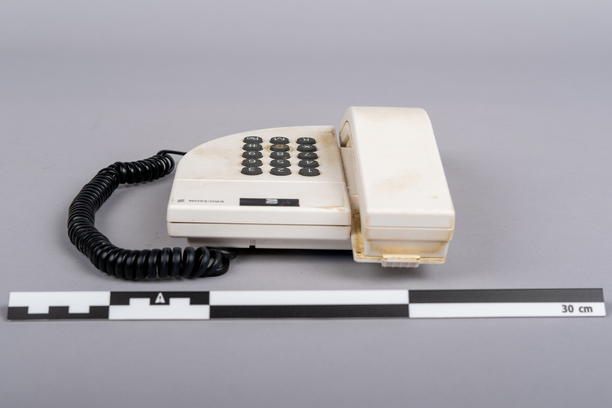 Ericsson bordtelefon.
Apparaten består av telefonrør, teelfon og spiralkabel. 
Hvit telefon med 15 svarte trykk knapper: tall (0-9), stjerne, nummertegn, funkjsonsknapper. 

Telefonen er misfarget, brune bruksmerker på telefonrør og rundt knappene, spesiell ved tallet 0.

På bunnen klistemerke med artikkel kode:
DBCN 2120700/987 R1A
ETM 9107 D. Design