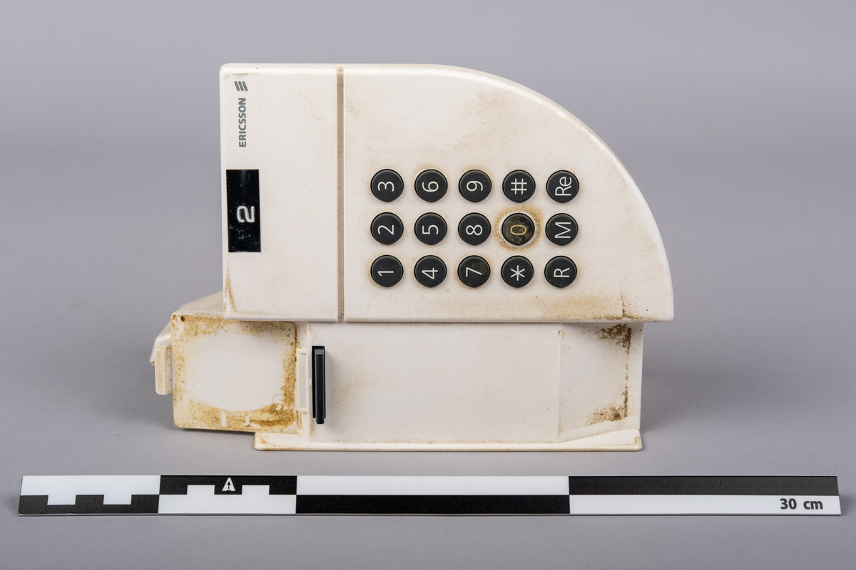 Ericsson bordtelefon.
Apparaten består av telefonrør, teelfon og spiralkabel. 
Hvit telefon med 15 svarte trykk knapper: tall (0-9), stjerne, nummertegn, funkjsonsknapper. 

Telefonen er misfarget, brune bruksmerker på telefonrør og rundt knappene, spesiell ved tallet 0.

På bunnen klistemerke med artikkel kode:
DBCN 2120700/987 R1A
ETM 9107 D. Design