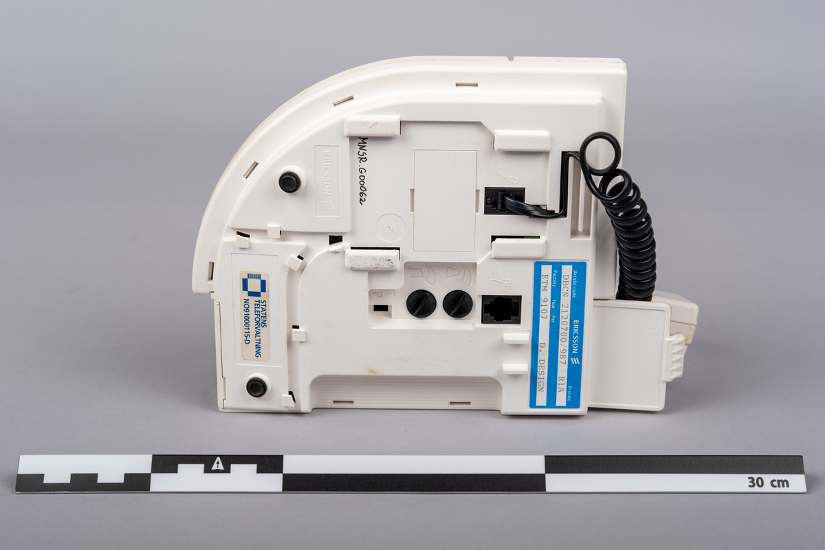 Ericsson bordtelefon.
Apparaten består av telefonrør, teelfon og spiralkabel. 
Hvit telefon med 15 svarte trykk knapper: tall (0-9), stjerne, nummertegn, funkjsonsknapper. 

Telefonen er misfarget, brune bruksmerker på telefonrør og rundt knappene, spesiell ved tallet 0.

På bunnen klistemerke med artikkel kode:
DBCN 2120700/987 R1A
ETM 9107 D. Design