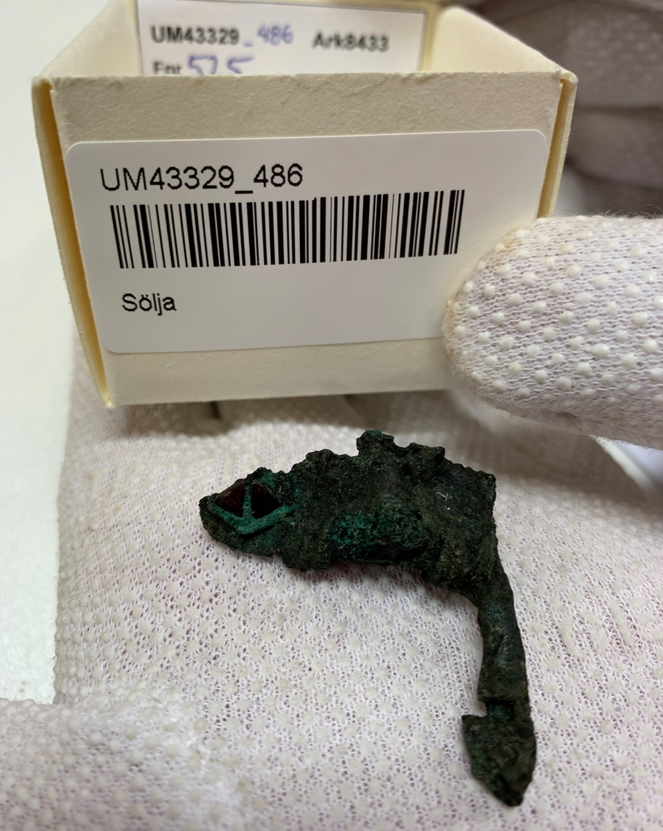 Föremål av kopparlegering. Sölja eller möjligen en remlöpare. Prydd med granater. Fragment.