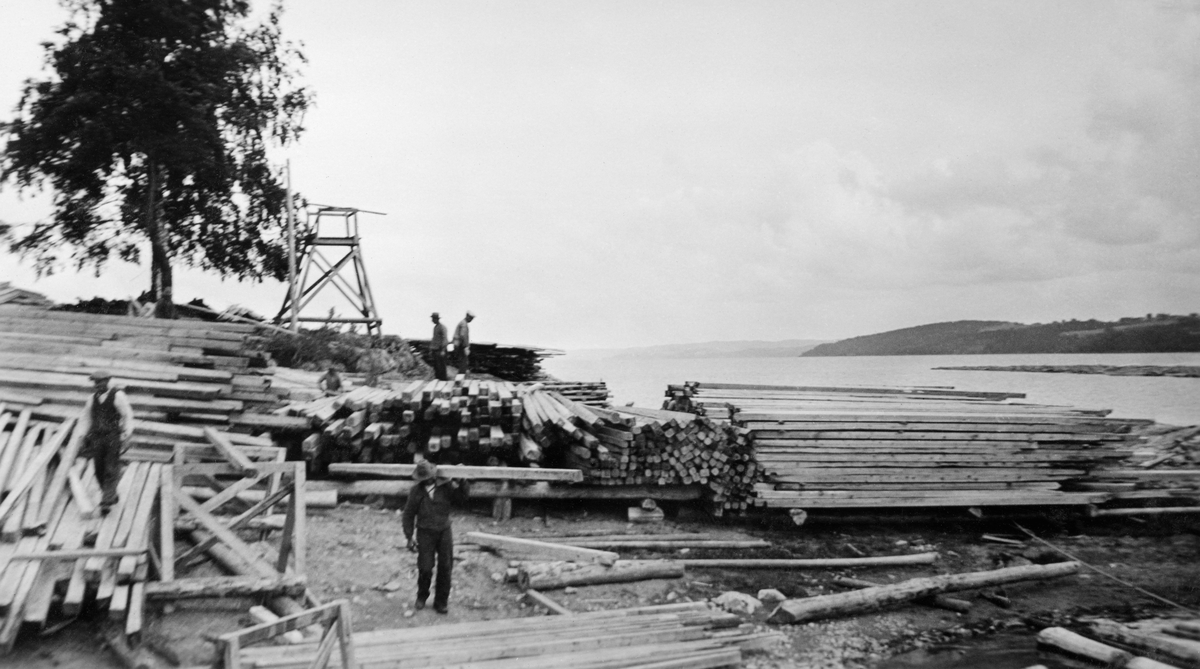 Materialstabler ved Sleppetangen i Spydeberg i Østfold i 1933. Da dette fotografiet ble tatt var fire menn i arbeid med å flytte på bjelker og planker. Vi vet foreløpig ikke hva disse materialene skulle brukes til. I bakgrunnen ser vi et tårnformet oppbygg som vi ikke kjenner funksjonen til.