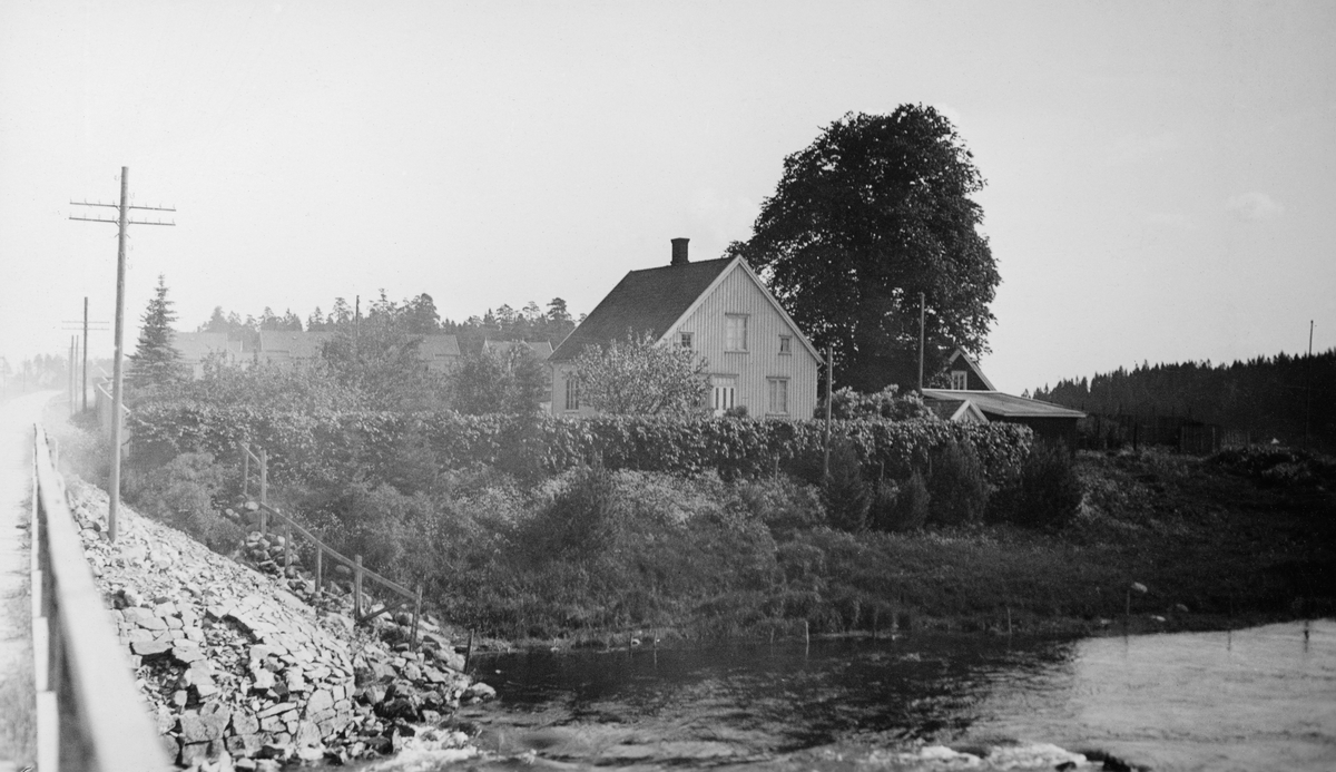 Fredrikstad Tømmerdirektions (Nedre Glommens fællesfløtningsforenings) damvokterbolig ved Sarpsborg i Østfold. Fotografiet later til å være tatt fra ei bru ved vassdraget mot er bordkledd halvannenetasjes våningshus med saltak og med dør på gavlveggen. Bakenfor skimter vi en noe mindre bygning, antakelig et uthus, og et flott tuntre. Mellom damvokterboligen og vassdraget var det en hekk.