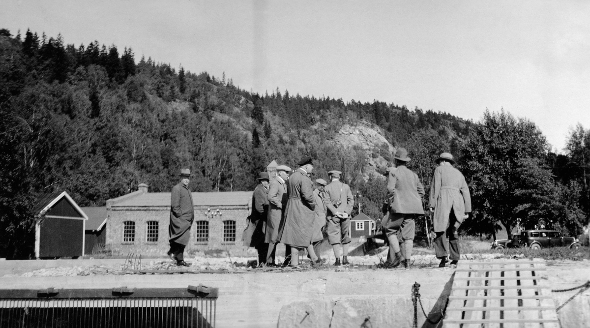 Vassdragskomiteén - antakelig Stortingets skog- og vassdragskomité - på befaring, antakelig ved Glomma fellesfløtingsforenings anlegg ved Eidet i Tune, der tømmertunnelen fra Isnesdfjorden munnet ut i Visterflo. Bildet er tatt mot en mur, der det sto cirka åtte menn i samtale. Bak dem ser vi et teglmurt maskinhus, der det sto en dampmaskin som antakelig drev de to mosemaskinene ved anlegget.