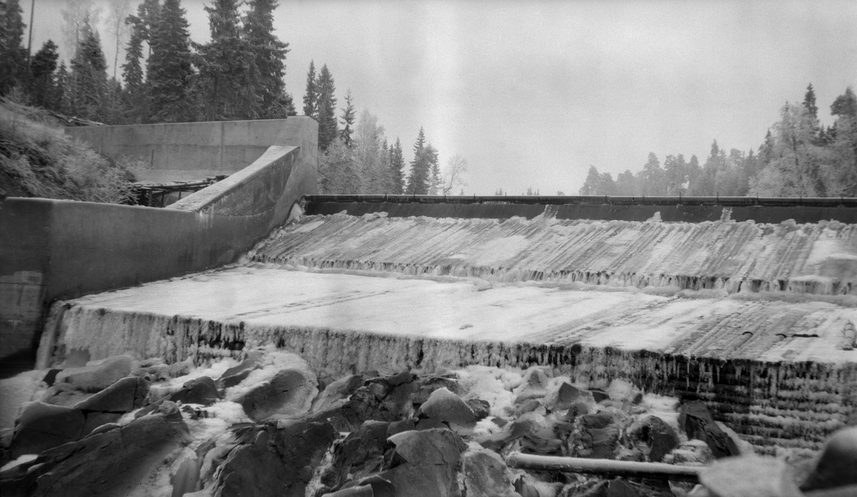 Nydammen i elva Søndre Osa i Åmot kommune i Østerdalen, fotografert i motstrøms retning i 1921. Dette var en terskel, støpt av betong og med sidevanger (skådammer) av betong. Nedenfor damkrona var det bygd et golv av tømmerstokker som lå tett og parallelt i strømretningen. Golvinga endte i en lav avsats mot det nedenforliggende elveløpet. Da dette fotografiet ble tatt var trekonstruksjonene dekt av et tynt islag og vannføringa var meget beskjeden.