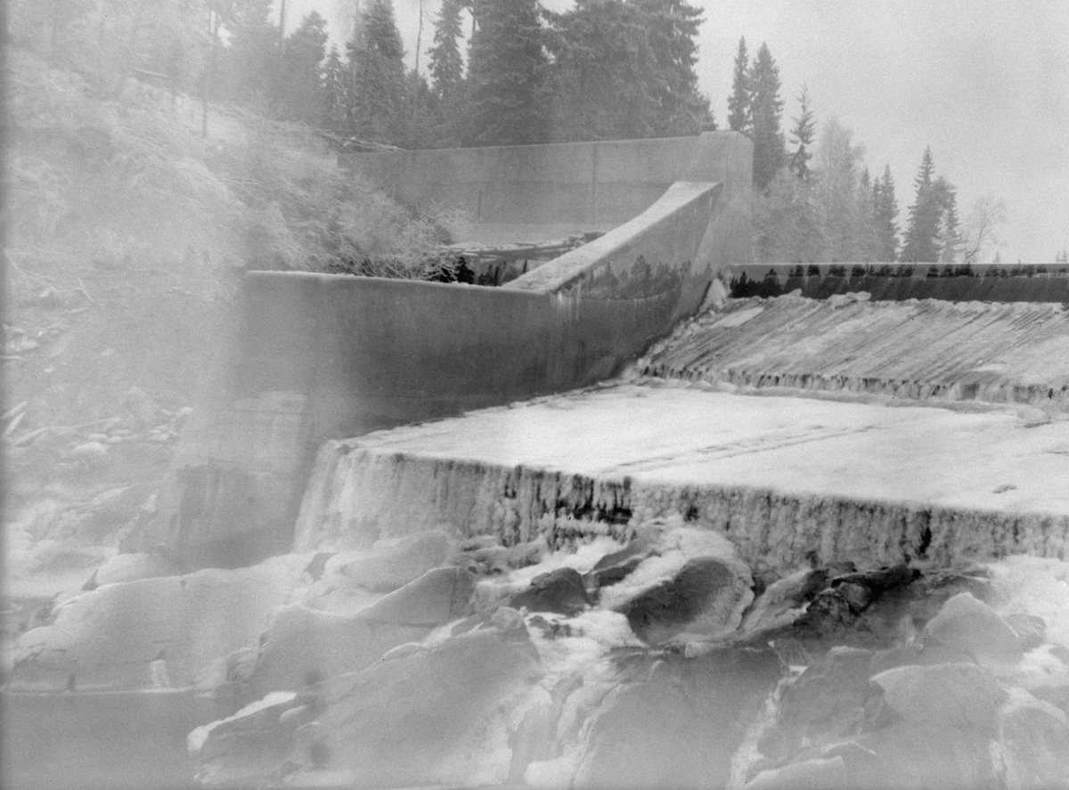 Nydammen i elva Søndre Osa i Åmot kommune i Østerdalen, fotografert i motstrøms retning i 1921. Dette var en terskel, støpt av betong og med sidevanger (skådammer) av betong. Nedenfor damkrona var det bygd et golv av tømmerstokker som lå tett og parallelt i strømretningen. Golvinga endte i en lav avsats mot det nedenforliggende elveløpet. Da dette fotografiet ble tatt var trekonstruksjonene dekt av et tynt islag og vannføringa var meget beskjeden.