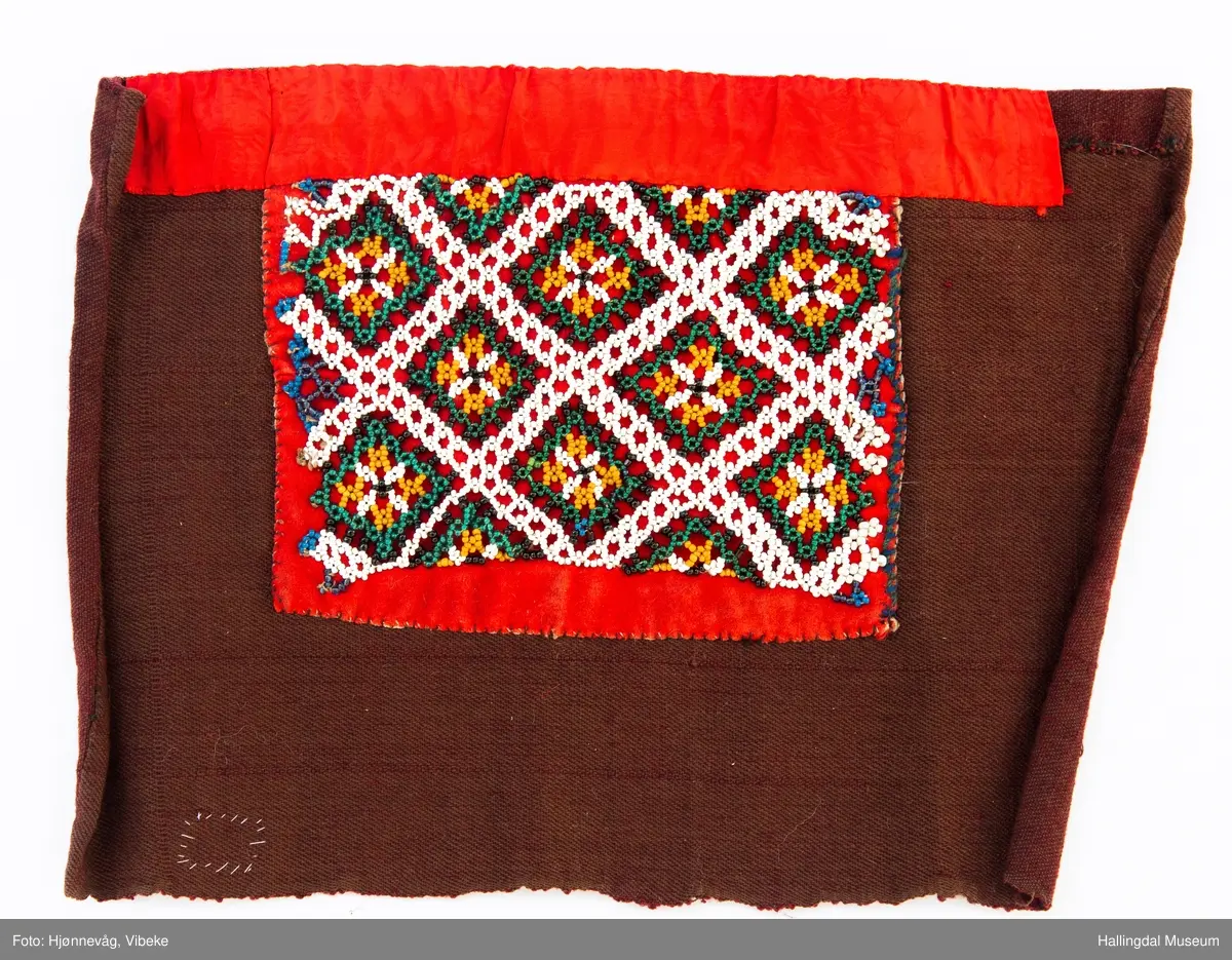 Perlet nett med små glassperler som er festet på et rødt klede. Hvite ruter med "blomster inni". Kledet er igjen festet på et brunt ullstoff. Øverst er et rødt silkebånd.