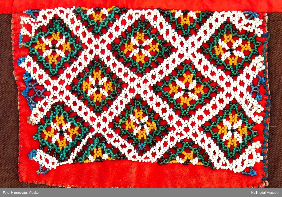 Perlet nett med små glassperler som er festet på et rødt klede. Hvite ruter med "blomster inni". Kledet er igjen festet på et brunt ullstoff. Øverst er et rødt silkebånd.