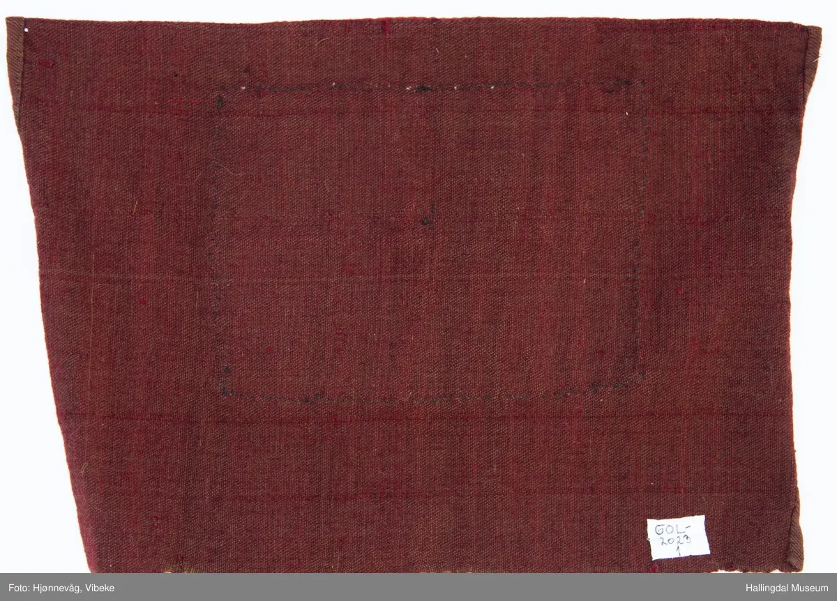 Perlet nett med små glassperler som er festet på et rødt klede. Hvite ruter med "blomster inni". Kledet er igjen festet på et brunt ullstoff. Øverst er et rødt silkebånd.