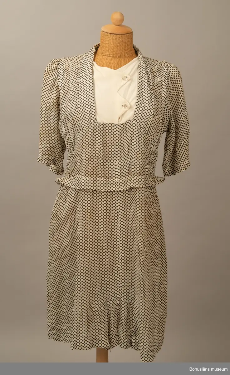 Hemmasydd i bomull (tuskaft med effektinslag). Vit botten med svart tryck. Små rutor på diagonalen. Djup fyrkantig urringning i halsen med vit insättning av muslin,som knäpps med två uddar och två glasknappar samt bendelband med tryckknapp. Avskuren på snedden upp mot ringningen. Lång krage fastsydd utmed ringningen och under avskärningen. Fyra sydda veck i midjan bak. Två nerstickade veck utmed ärmens bak och framstycke. Ett veck innanför detta. Fyra infällda veck på kjol-vådens nederkant fram. Kort ärm med dragsko.

Sliten och blekt. Många lagningar.