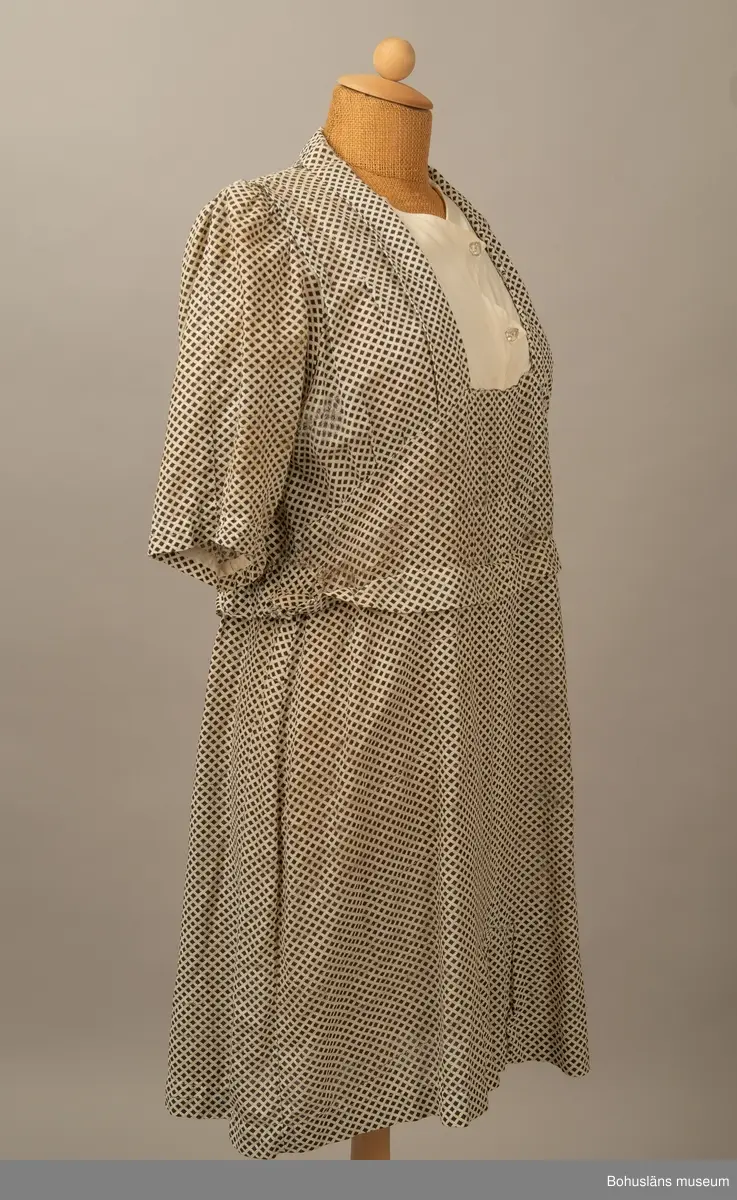 Hemmasydd i bomull (tuskaft med effektinslag). Vit botten med svart tryck. Små rutor på diagonalen. Djup fyrkantig urringning i halsen med vit insättning av muslin,som knäpps med två uddar och två glasknappar samt bendelband med tryckknapp. Avskuren på snedden upp mot ringningen. Lång krage fastsydd utmed ringningen och under avskärningen. Fyra sydda veck i midjan bak. Två nerstickade veck utmed ärmens bak och framstycke. Ett veck innanför detta. Fyra infällda veck på kjol-vådens nederkant fram. Kort ärm med dragsko.

Sliten och blekt. Många lagningar.