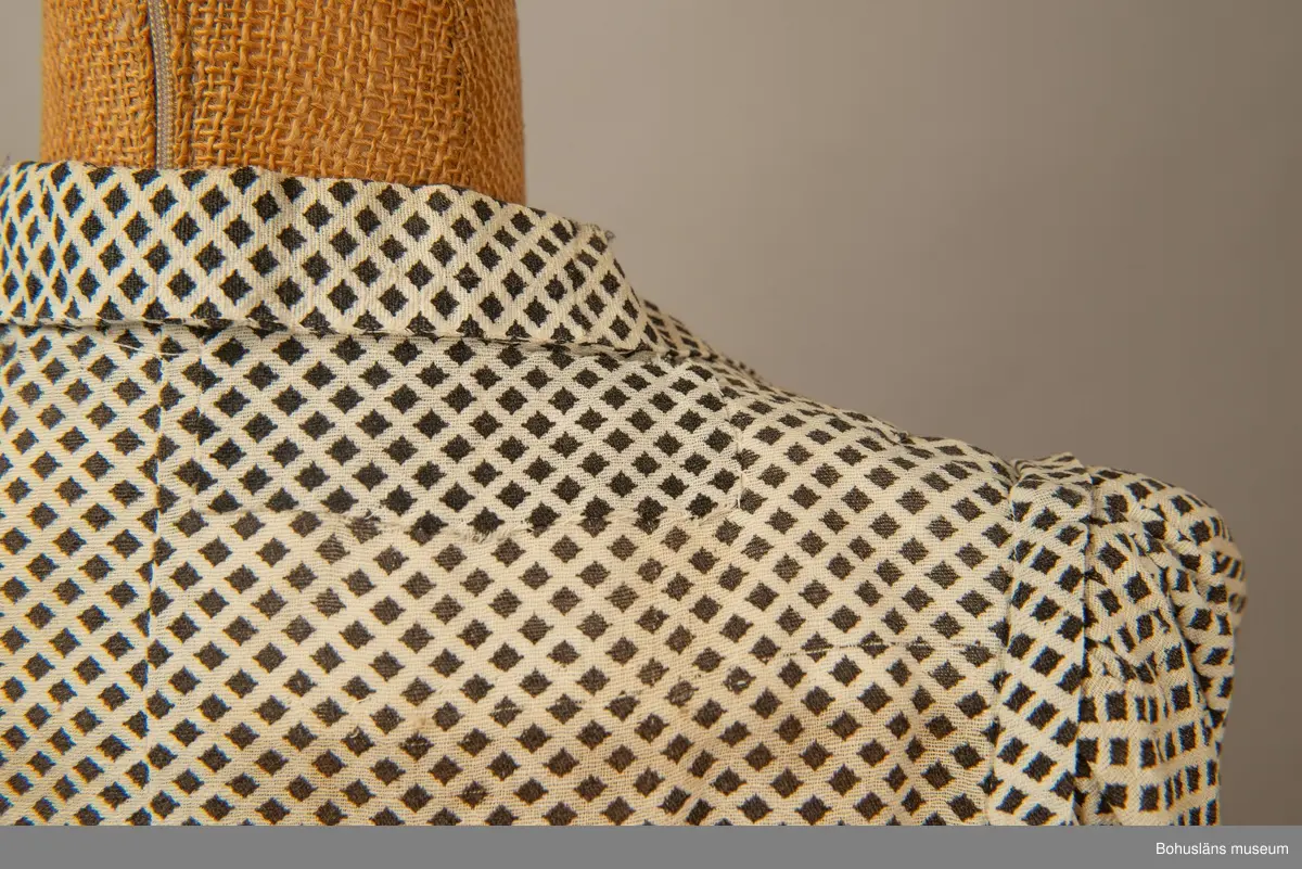 Hemmasydd i bomull (tuskaft med effektinslag). Vit botten med svart tryck. Små rutor på diagonalen. Djup fyrkantig urringning i halsen med vit insättning av muslin,som knäpps med två uddar och två glasknappar samt bendelband med tryckknapp. Avskuren på snedden upp mot ringningen. Lång krage fastsydd utmed ringningen och under avskärningen. Fyra sydda veck i midjan bak. Två nerstickade veck utmed ärmens bak och framstycke. Ett veck innanför detta. Fyra infällda veck på kjol-vådens nederkant fram. Kort ärm med dragsko.

Sliten och blekt. Många lagningar.