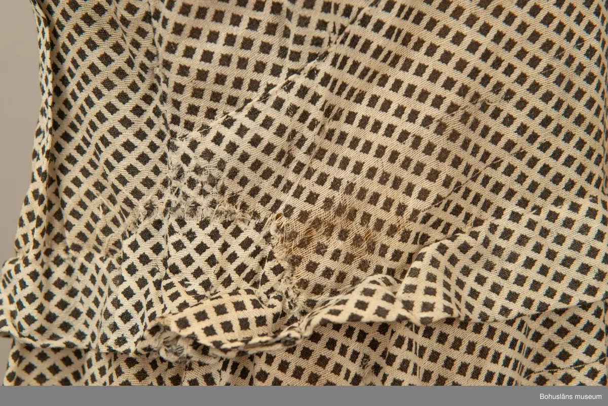 Hemmasydd i bomull (tuskaft med effektinslag). Vit botten med svart tryck. Små rutor på diagonalen. Djup fyrkantig urringning i halsen med vit insättning av muslin,som knäpps med två uddar och två glasknappar samt bendelband med tryckknapp. Avskuren på snedden upp mot ringningen. Lång krage fastsydd utmed ringningen och under avskärningen. Fyra sydda veck i midjan bak. Två nerstickade veck utmed ärmens bak och framstycke. Ett veck innanför detta. Fyra infällda veck på kjol-vådens nederkant fram. Kort ärm med dragsko.

Sliten och blekt. Många lagningar.