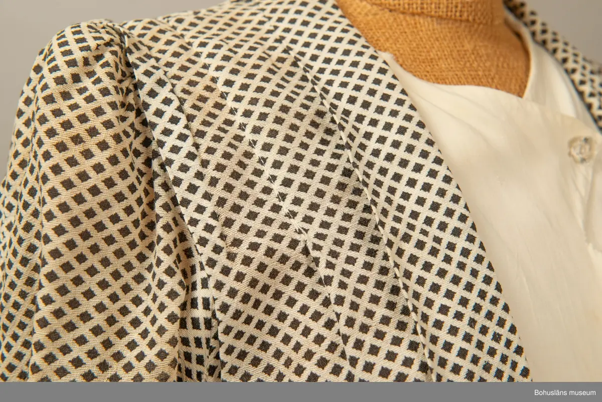 Hemmasydd i bomull (tuskaft med effektinslag). Vit botten med svart tryck. Små rutor på diagonalen. Djup fyrkantig urringning i halsen med vit insättning av muslin,som knäpps med två uddar och två glasknappar samt bendelband med tryckknapp. Avskuren på snedden upp mot ringningen. Lång krage fastsydd utmed ringningen och under avskärningen. Fyra sydda veck i midjan bak. Två nerstickade veck utmed ärmens bak och framstycke. Ett veck innanför detta. Fyra infällda veck på kjol-vådens nederkant fram. Kort ärm med dragsko.

Sliten och blekt. Många lagningar.