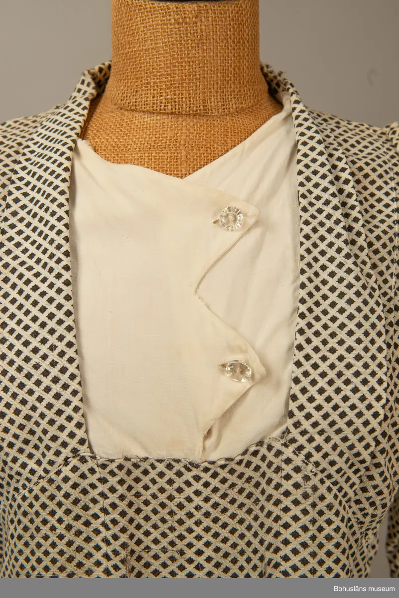 Hemmasydd i bomull (tuskaft med effektinslag). Vit botten med svart tryck. Små rutor på diagonalen. Djup fyrkantig urringning i halsen med vit insättning av muslin,som knäpps med två uddar och två glasknappar samt bendelband med tryckknapp. Avskuren på snedden upp mot ringningen. Lång krage fastsydd utmed ringningen och under avskärningen. Fyra sydda veck i midjan bak. Två nerstickade veck utmed ärmens bak och framstycke. Ett veck innanför detta. Fyra infällda veck på kjol-vådens nederkant fram. Kort ärm med dragsko.

Sliten och blekt. Många lagningar.