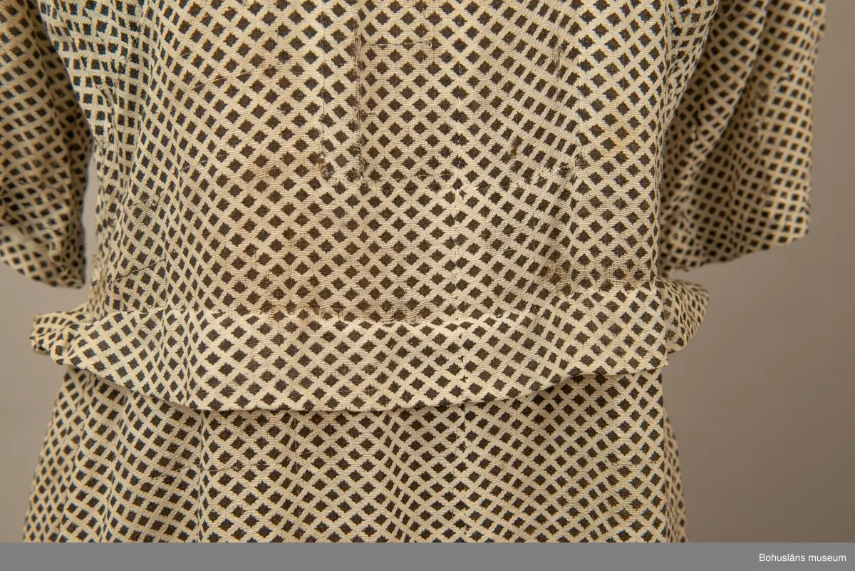 Hemmasydd i bomull (tuskaft med effektinslag). Vit botten med svart tryck. Små rutor på diagonalen. Djup fyrkantig urringning i halsen med vit insättning av muslin,som knäpps med två uddar och två glasknappar samt bendelband med tryckknapp. Avskuren på snedden upp mot ringningen. Lång krage fastsydd utmed ringningen och under avskärningen. Fyra sydda veck i midjan bak. Två nerstickade veck utmed ärmens bak och framstycke. Ett veck innanför detta. Fyra infällda veck på kjol-vådens nederkant fram. Kort ärm med dragsko.

Sliten och blekt. Många lagningar.