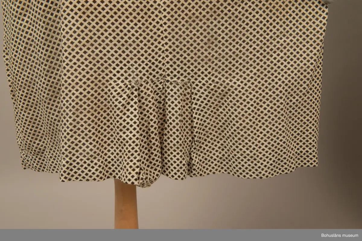 Hemmasydd i bomull (tuskaft med effektinslag). Vit botten med svart tryck. Små rutor på diagonalen. Djup fyrkantig urringning i halsen med vit insättning av muslin,som knäpps med två uddar och två glasknappar samt bendelband med tryckknapp. Avskuren på snedden upp mot ringningen. Lång krage fastsydd utmed ringningen och under avskärningen. Fyra sydda veck i midjan bak. Två nerstickade veck utmed ärmens bak och framstycke. Ett veck innanför detta. Fyra infällda veck på kjol-vådens nederkant fram. Kort ärm med dragsko.

Sliten och blekt. Många lagningar.