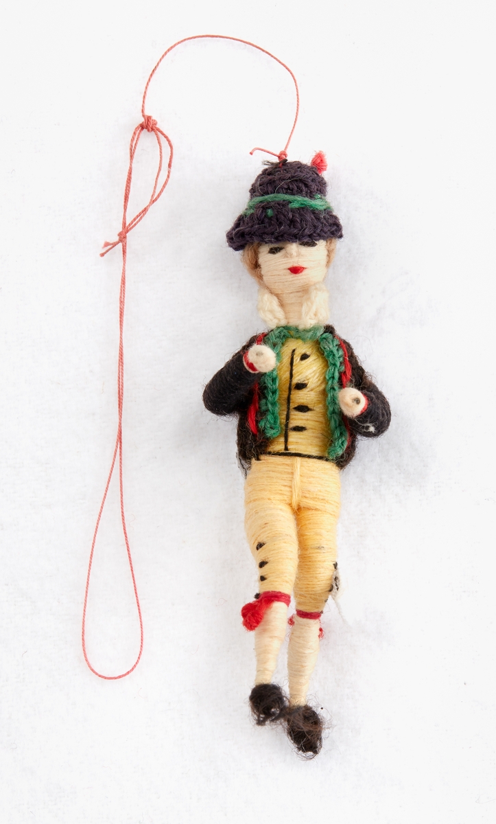Docka i landskaps/sockendräkt. Dockans kläder är av tråd lindat runt en stomme av metall. Dockan har gula byxor, svart jacka med gröna kanter och detaljer i rosa, svart hatt med grön kant, röda knätofsar, svarta skor.

Från donationen Garpe-Hedestrand 2000.

Höjd: 90 mm.
Bredd: 30 mm.
