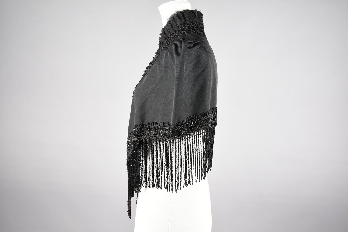 Cape. Perlebroderi og lissebroderi. Bord rundt hals, foran og rundt livet. Blonderysjer og hals foran. Skuldrene er markert med legg. Hekter fremme. Perlebrodert, lange perlefrynser.