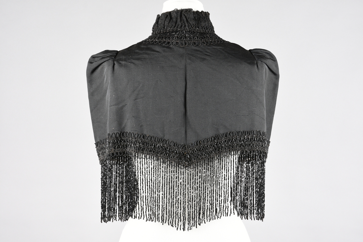 Cape. Perlebroderi og lissebroderi. Bord rundt hals, foran og rundt livet. Blonderysjer og hals foran. Skuldrene er markert med legg. Hekter fremme. Perlebrodert, lange perlefrynser.