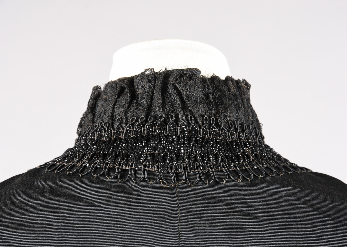 Cape. Perlebroderi og lissebroderi. Bord rundt hals, foran og rundt livet. Blonderysjer og hals foran. Skuldrene er markert med legg. Hekter fremme. Perlebrodert, lange perlefrynser.