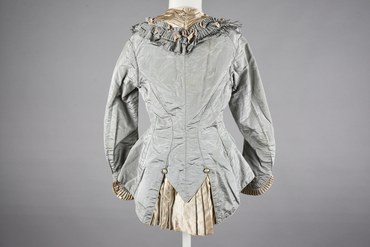 Liv av lysgrå taftsilke. Knappet foran. Rysjekrave. Figursydd. Kraven er et stykke legget med lysere grå og blank silke. Det samme er plissert i skjøten bak. Beltebånd innvendig, 14 cm. Trukne knapper. Trange ermer med rysj nede.
Merket med blekk innvendig i beltebåndet: Grung.