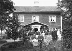 Sällskap samlat för fotografering framför Hageby Östergård i Skedevi. Enligt påskrift 1890-tal, i tid då gården ägdes och brukades av makarna August och Anna Jonsson. Möjligtvis är det dem vi ser sittande centralt i bilden.