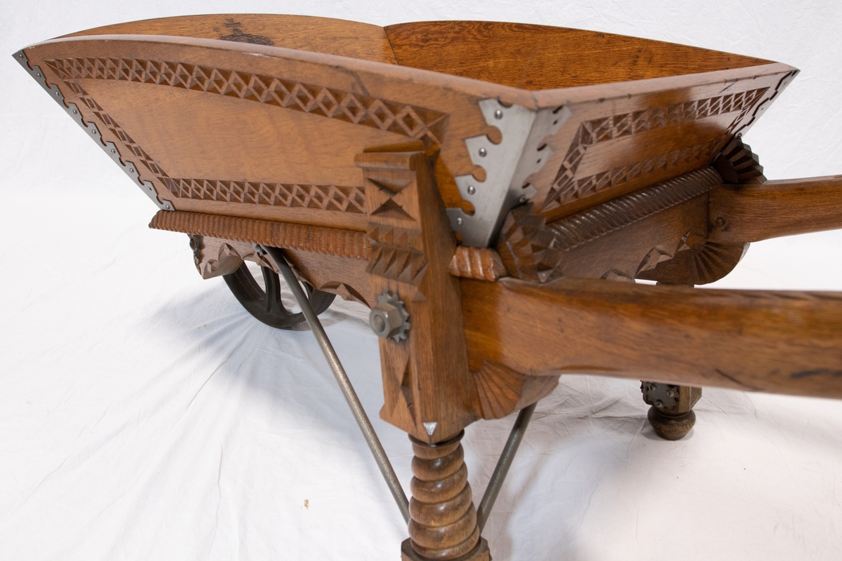Trillebår av tre med metalhjul og detaljer. Trillebåren er dekorativt ingravert med blant annet drage detaljer.