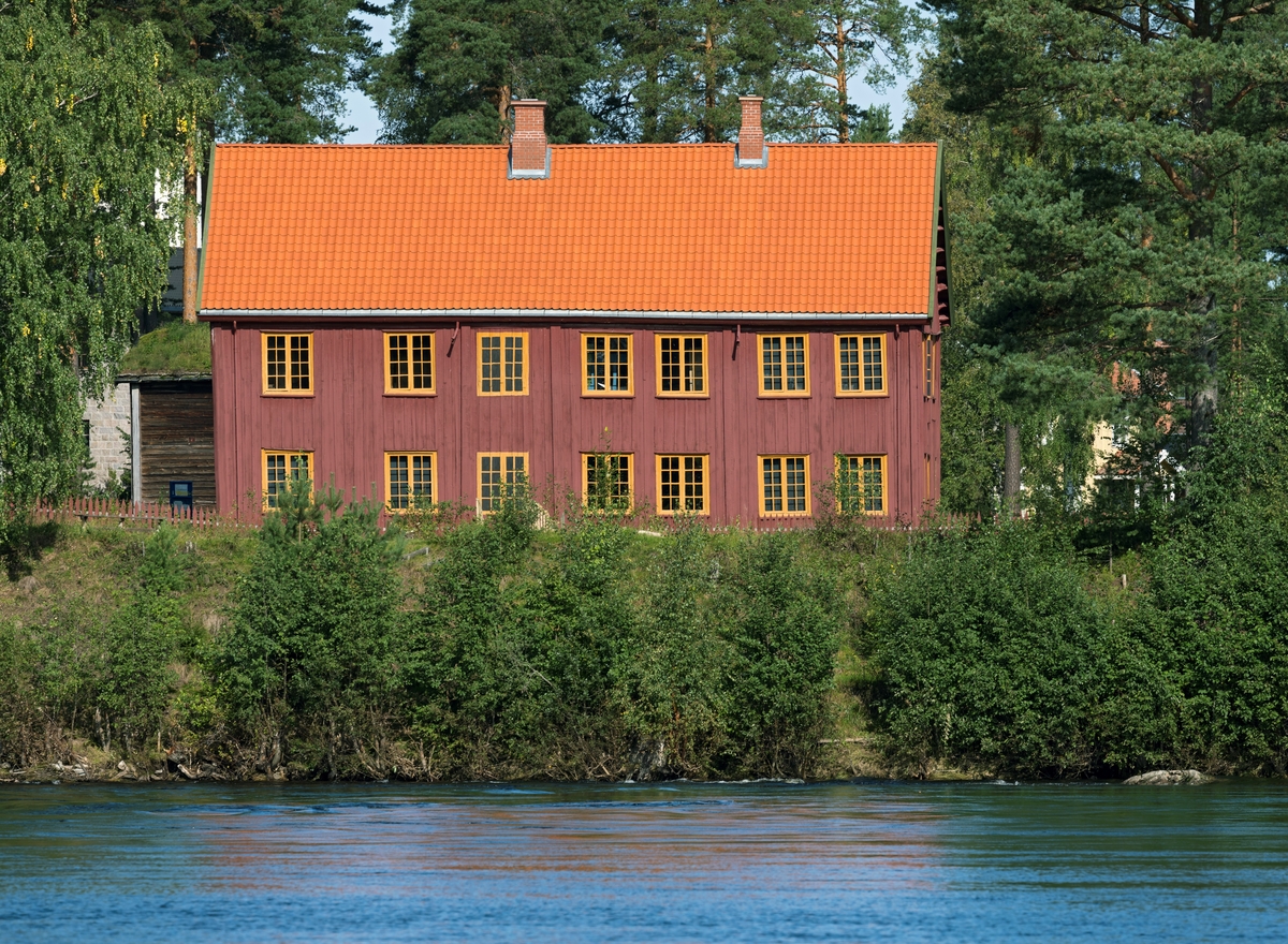 Åset-bygningen på Glomdalsmuseet i Elverum. Dette var opprinnelig ett av våningshusa på en stor jord- og skogeiendom i Åmot i Østerdalen. Glomdalsmuseet ble etablert som et friluftsmuseum med tilflyttete bygninger fra bygdene i Solør og Østerdalen. Den første av disse tilflyttete bygningene, ei barfrøstue fra Rendalen, ble innkjøpt i 1910. Da museet feiret sitt 15-årsjubileum i 1926 besto museet av 30 bygninger, og 9 ventet på flytting til museumsparken på Vestad i Elverum. Det digre våningshuset på Nordre Åset i Åmot var ett av disse. Åset-bygningen ble bygd i 1795, mens Ole Olsen (1767-1825) og Anne Tollevsdatter (1772-1844) drev garden. Konservator Håvard Skirbekk (1903-1983) skrev at «Åsetbygningen er bygd i 1795, og skal ha vori den første tohøgda bygningen i Åmot. Det var ikkje så mange i Elverum heller før den tid. Men 30 år seinare var det mange storgardsbygningar av liknande type.» Åset-bygningen er 18 meter lang og 8,8 meter bred, hadde to fulle etasjer og et forholdsvis spissrøstet saltak, så den ruvde godt i tunet på Nordre Åset. Det gjorde den for så vidt etter at den ble flyttet til Glomdalsmuseet også. Den 6. juni 1927 kunne avisa «Østlændingen» melde at «Åmot-anlegget er nu påbegynt, idet den store hovedbygning på Nordre Åset blev revet ned og fraktet til museet i vinter. Bygningen vil komme under tak i løpet av vinteren.» Den 31. mai 1928 meldte den samme avisa at bygningen sto ferdig gjenreist på museet. Gjenoppføringa gikk altså forholdsvis raskt unna. Etter hvert skulle det vise seg at det hadde gått litt for fort. Museet ligger på en furumo med sandgrunn, og akkurat der Åset-bygningen ble plassert skal det ha vært en litt fuktig forsenkning. Mangelfullt grunnarbeid førte til at grunnen gav etter for vekta av de tunge bygningskonstruksjonene, som seg stadig lengre ned i bakken. Det var for øvrig ikke alle deler av huset som sank like mye, noe som også skapte vedlikeholdsutfordringer. Bortimot 90 år etter at Åset-bygningen ble gjenreist på Glomdalsmuseet ble det derfor igangsatt et stort vedlikeholdsprosjekt som gikk over mange sesonger og kostet store pengebeløp. Dette fotografiet er tatt i 2023, et par år etter at prosjektet var avsluttet. Her ser vi det som på museet ble østre langvegg, som er rødmalt med gule vindusomramminger. Taket hadde fått nytt tekke av krumtegl. Opptaket er gjort med telelinse over Glomma på et tidspunkt med regnflom og unormalt høy vannstand i elva.
