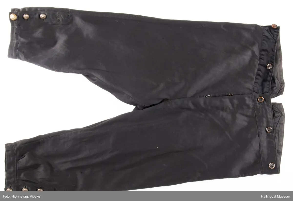 Svart knebukse i silke/bomull, glinsende stoff. 3 sølvknapper på hver fot, 4 håndsydde knapphull. Knapper i gylfen og til bukseseler på linningen. Spensel i ryggen.