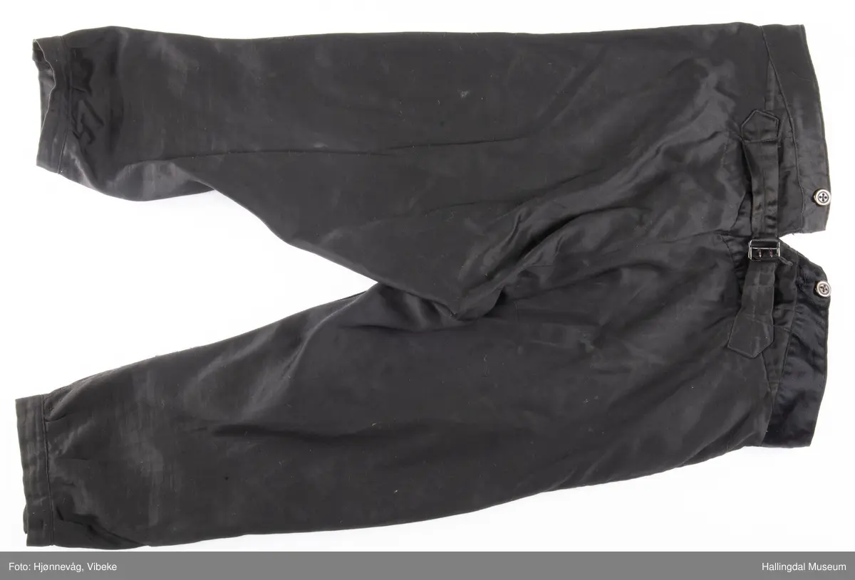 Svart knebukse i silke/bomull, glinsende stoff. 3 sølvknapper på hver fot, 4 håndsydde knapphull. Knapper i gylfen og til bukseseler på linningen. Spensel i ryggen.