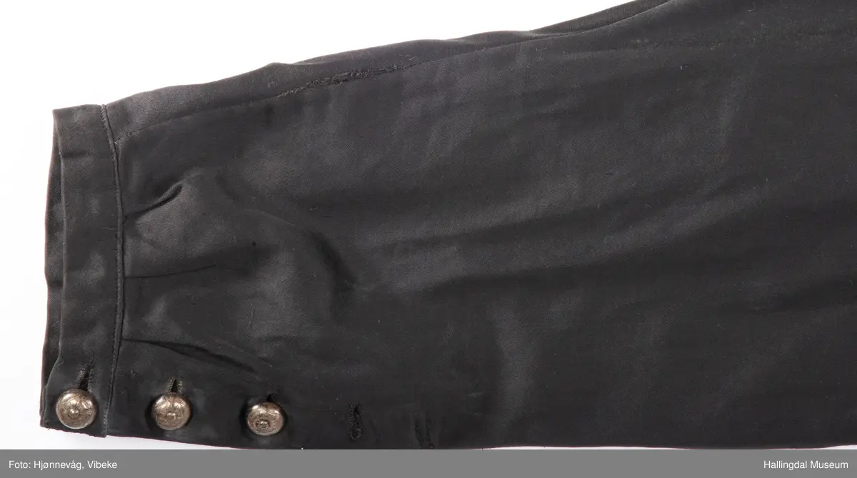 Svart knebukse i silke/bomull, glinsende stoff. 3 sølvknapper på hver fot, 4 håndsydde knapphull. Knapper i gylfen og til bukseseler på linningen. Spensel i ryggen.