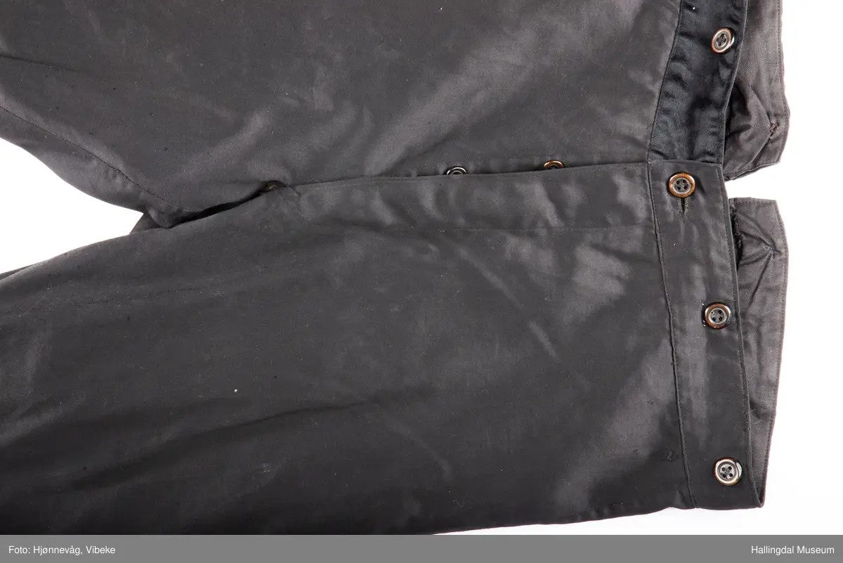 Svart knebukse i silke/bomull, glinsende stoff. 3 sølvknapper på hver fot, 4 håndsydde knapphull. Knapper i gylfen og til bukseseler på linningen. Spensel i ryggen.